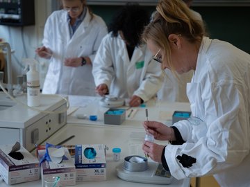 Drei Frauen in Laborkitteln wiegen chemische Stoffe mit Waagen ab.