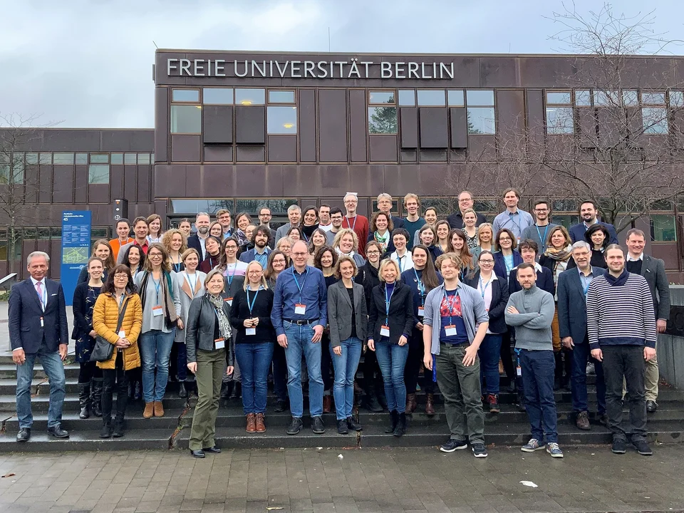 Eine Gruppe von Personen steht vor einem Gebäude der Freien Universität Berlin auf einer Treppe, einige tragen Namensschilder.