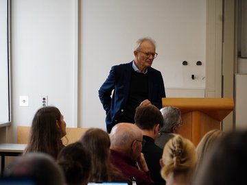 Jonathan Osborne bei der AQUA-d Konferenz. Er steht neben dem Pult; im Vordergrund sind die Hinterköpfe von Zuhörende zu sehen. 