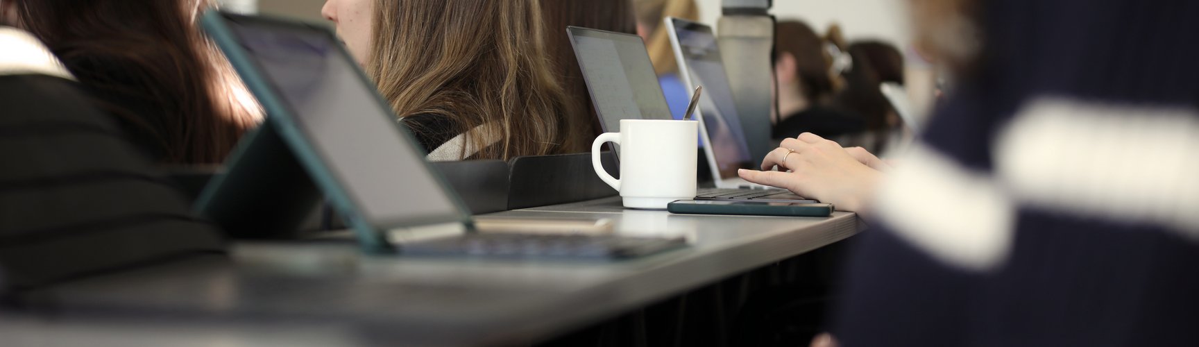 Szene aus einem Hörsaal, im Zentrum steht eine Kaffeetasse