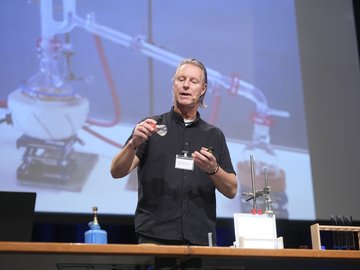 Ein Vortragender steht auf dem Podium, vor ihm chemische Laborgeräte, hinter ihm ist eine Präsentation auf der Leinwand zu sehen.