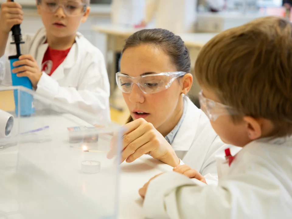 Eine Studentin macht zusammen mit einem Grundschulkind ein chemisches Experiment. Beide tragen Schutzbrillen.
