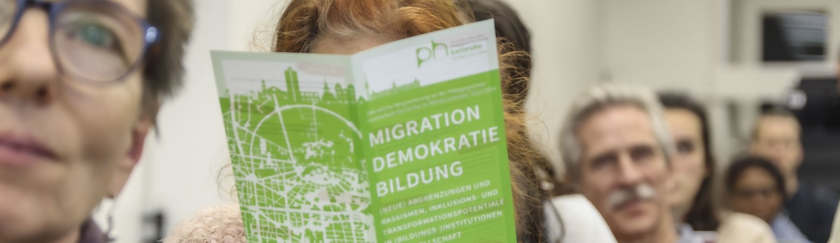 Im Publikum hebt jemand einen Flyer mit dem Titel "Migration Bildung Demokratie" hoch