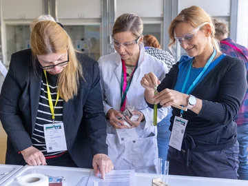 Drei Frauen betrachten gemeinsam Unterlagen und chemische Laborgeräte. Eine davon trägt einen Laborkittel.