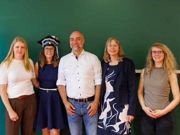 5 freundliche Personen vor einer grünen Tafel. Eine Person trägt einen verzierten Doktorhut.