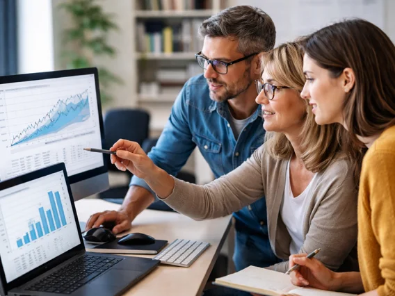 Zwei Frauen und ein Mann schauen auf einen Desktop- und einen Laptop-Bildschirm, die Schaubilder analytischer Daten zeigen. Sie sprechen darüber.