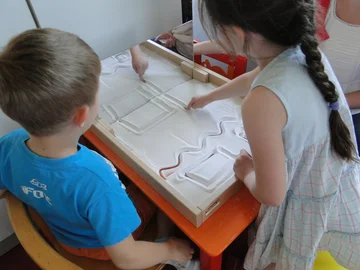 Zwei Kinder zeichnen mit dem Finger Muster in einer Wanne voller Sand.