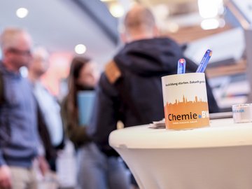 Nahaufnahme einer Tasse mit Chemie-BW-Logo. Menschen im Hintergrund.