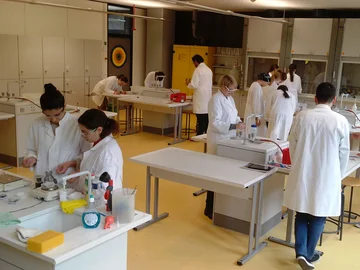 In einem Labor stehen mehrere Menschen mit Laborkitteln und Schutzbrillen an Labortischen und arbeiten an Versuchsaufbauten.
