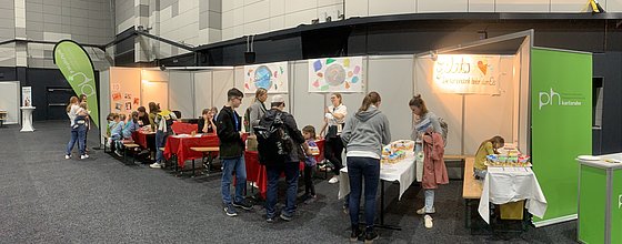 Reihe der Ausstellertische der PHKA bei den Science DAys für Kinder mit roten Tischdecken. Flankiert wird es von aufstelern mit dem PHKA Logo weiß auf Grün. Um die Tische herum sitzen und stehen KInder und Studierende.