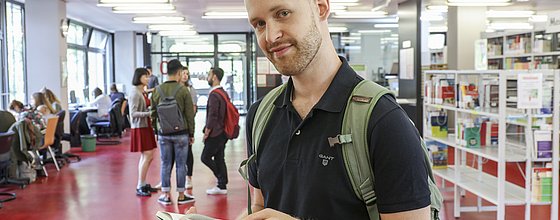 Ein Student, von vorne fotografiert, liest ein Buch in der Bibliothek im Gebäude 2 der PH Karlsruhe. Hinter ihm sind mehrere Studierende zu sehen. ©PHKA