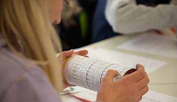 LLL_Informatik_Abstraktion Eien Schülerin von hinten, die konzentriert auf bildlich dargestellte Programmierschritte auf einer Rolle blickt.