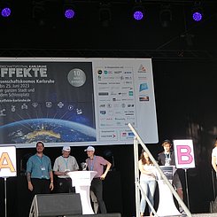 Moderator und Kandidat:innen des Quiz "Geistesblitze - Duell der klugen Köpfe" auf der EFFEKTE-Event-Bühne. Es sind die Stationen A und B zu sehen. Rechts vom A steht Jun.Prof. Dr. Sebastian Engelmann, rechts vom B eine PHKA Lehramtsstudentin. Im Hintergrund ist das EFFEKTE Bahner aufgespannt mit dem Plakatmotiv und den Logos aller beteiligten Institutionen.