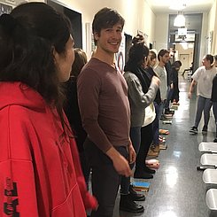 Auf dem Bild ist ein Stundeneinstieg zu sehen.Studierende stehen in einer Reihe zwischen Bücherstapeln.